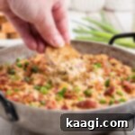 A golden brown skillet filled with creamy Boudin Dip, garnished with fresh green onions, with a hand dipping a chip into the dip.
