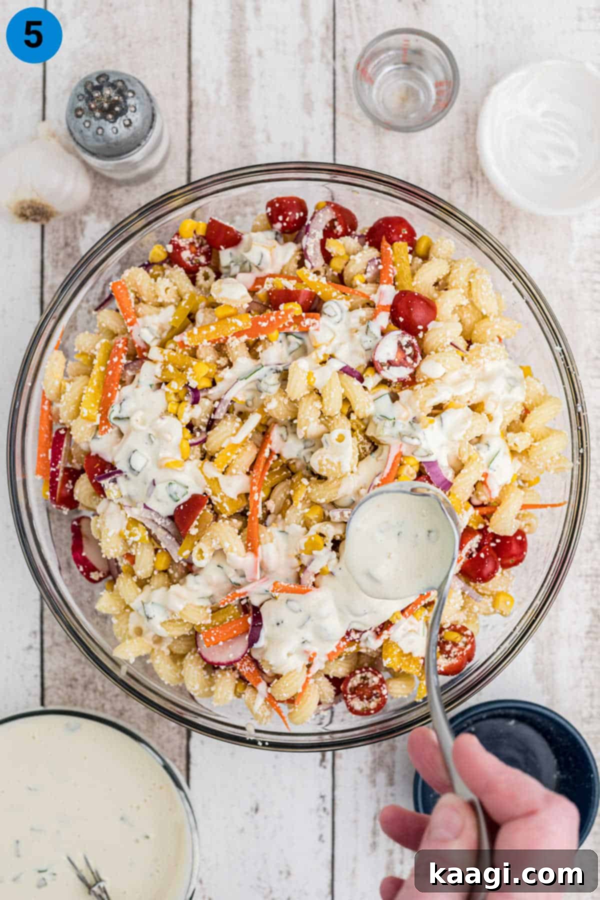 Creamy white dressing being generously poured over a colorful bowl of pasta salad, ready to be mixed.