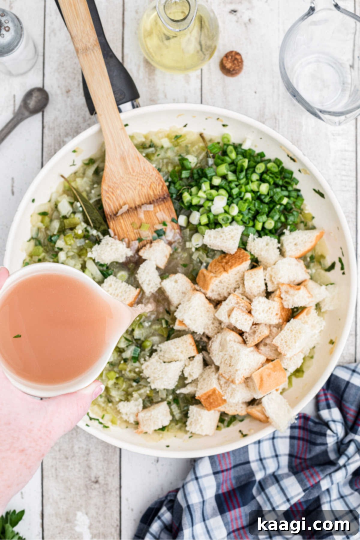 Bread cubes, chopped green onions, and reserved oyster liquor being added to the skillet with cooked vegetables.