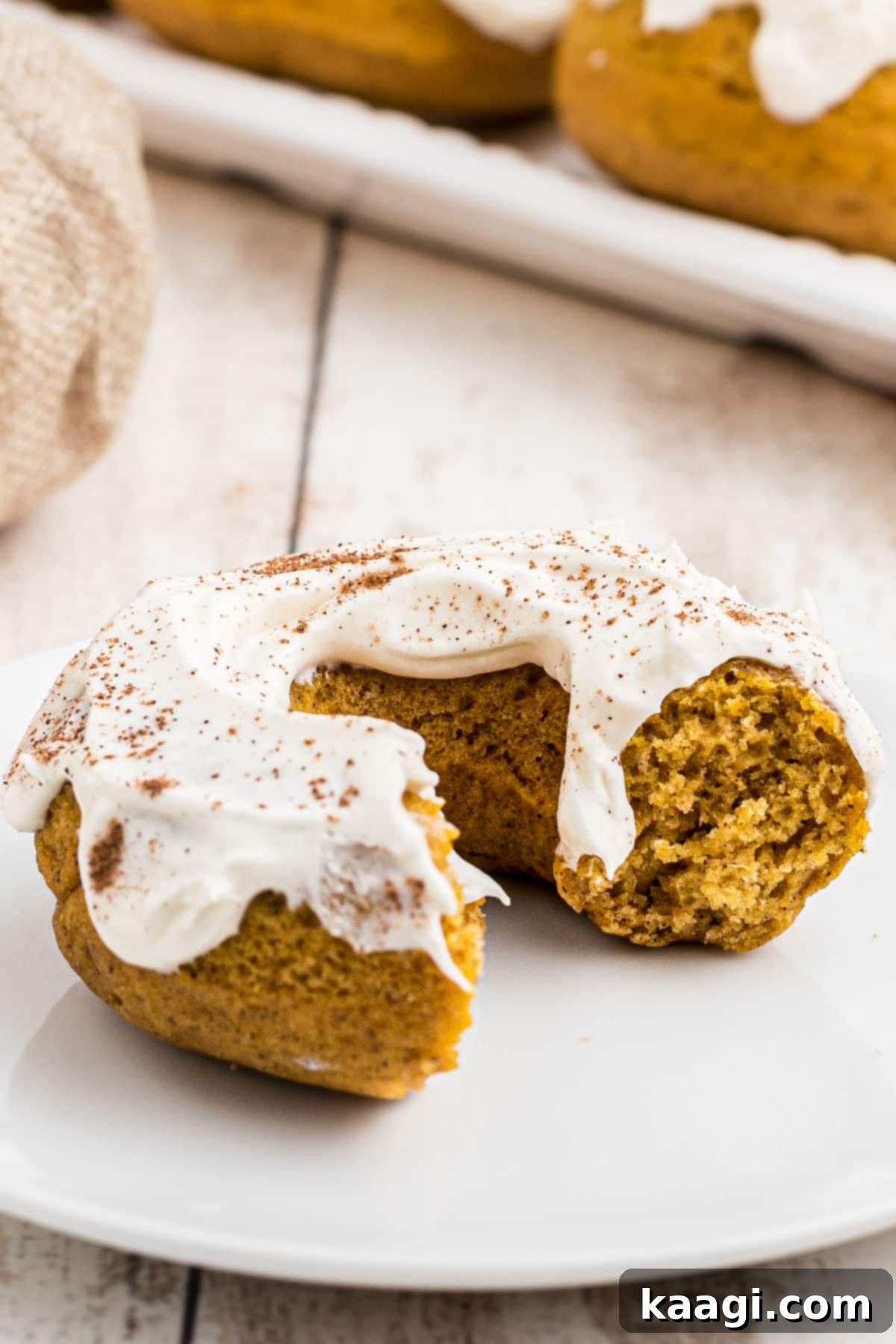 Close-up of a perfectly baked pumpkin spice donut with a bite taken out, showcasing the fluffy interior and thick cream cheese frosting, with more donuts blurred in the background.