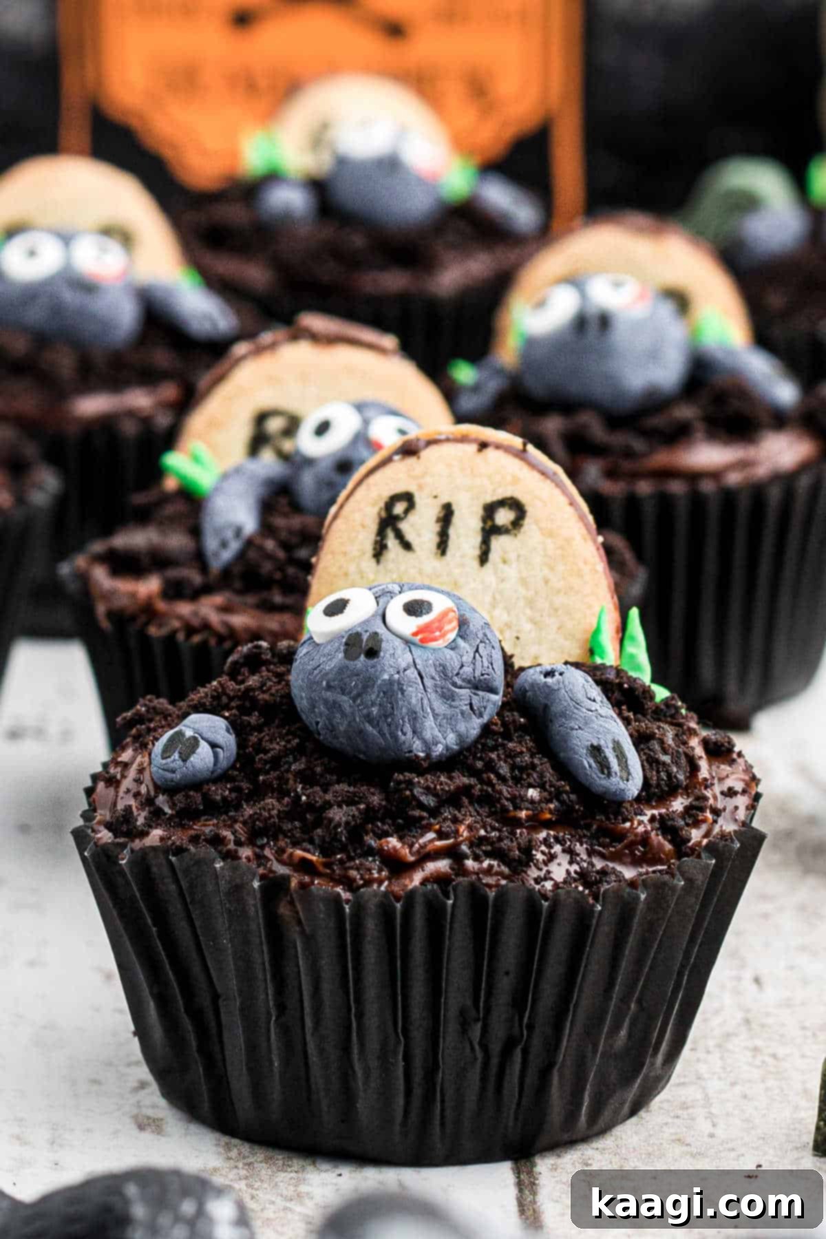 Delicious Halloween Zombie Cupcakes Close up of a zombie cupcake with more zombie cupcakes behind, showcasing the detailed fondant zombie, crushed Oreo 'earth', and an oval cookie 'tombstone'.