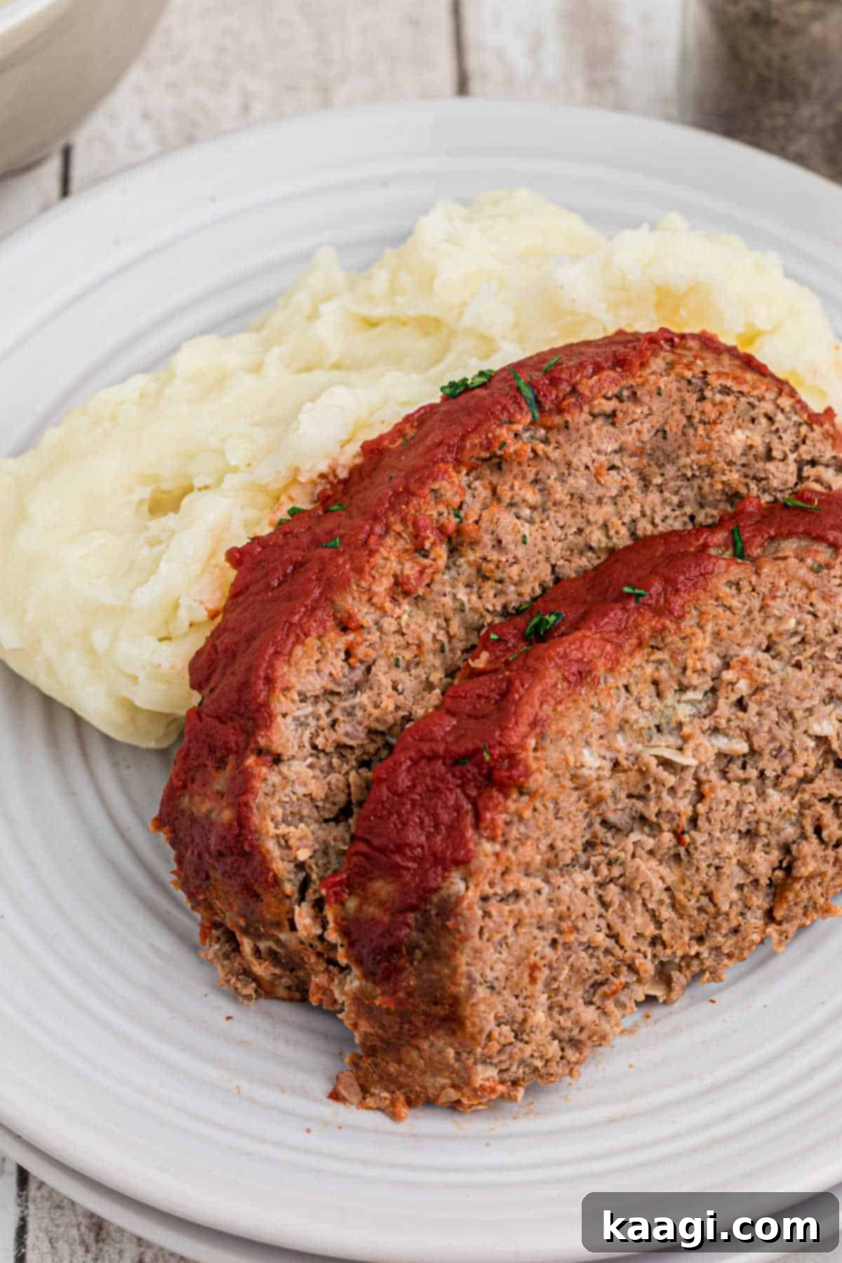 Two generous slices of perfectly cooked venison meatloaf, glazed with tomato sauce and nestled beside a mound of creamy mashed potatoes, ready to be enjoyed.