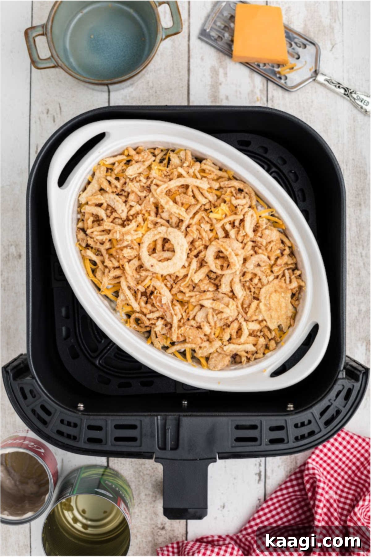 An air fryer safe casserole dish topped with grated cheddar cheese and crispy French fried onions, positioned inside an air fryer basket, ready to be cooked.