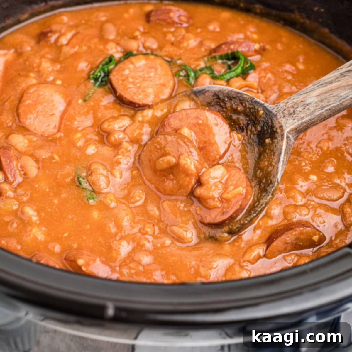 close up of kielbasa and baked beans with a wooden spoon taking some.