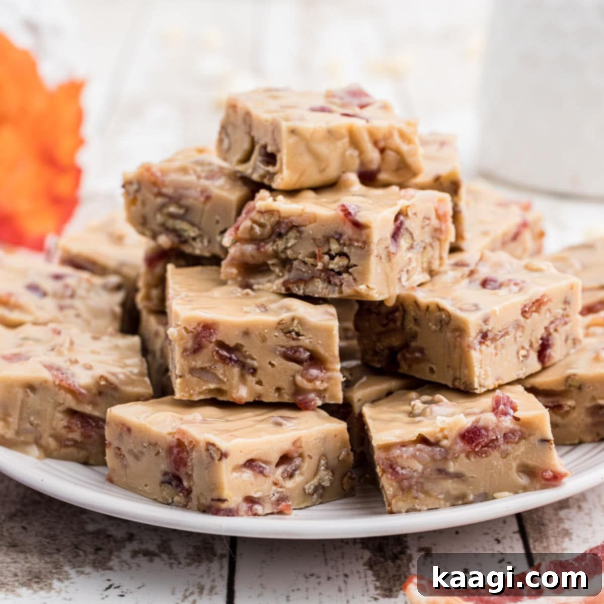 A plate generously filled with perfectly cut squares of maple bacon fudge, with a decorative maple leaf and crispy bacon slices artfully arranged in the soft-focus background.