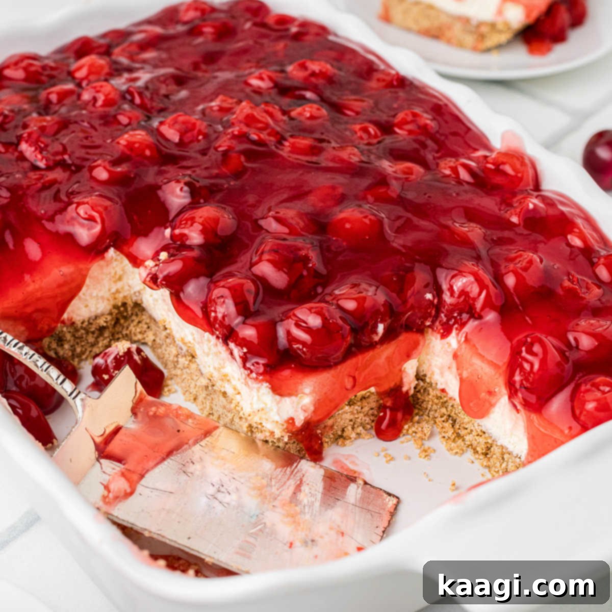 A large glass baking dish filled with vibrant old-fashioned cherry delight, with a few squares already taken and a serving spatula resting inside the dish.