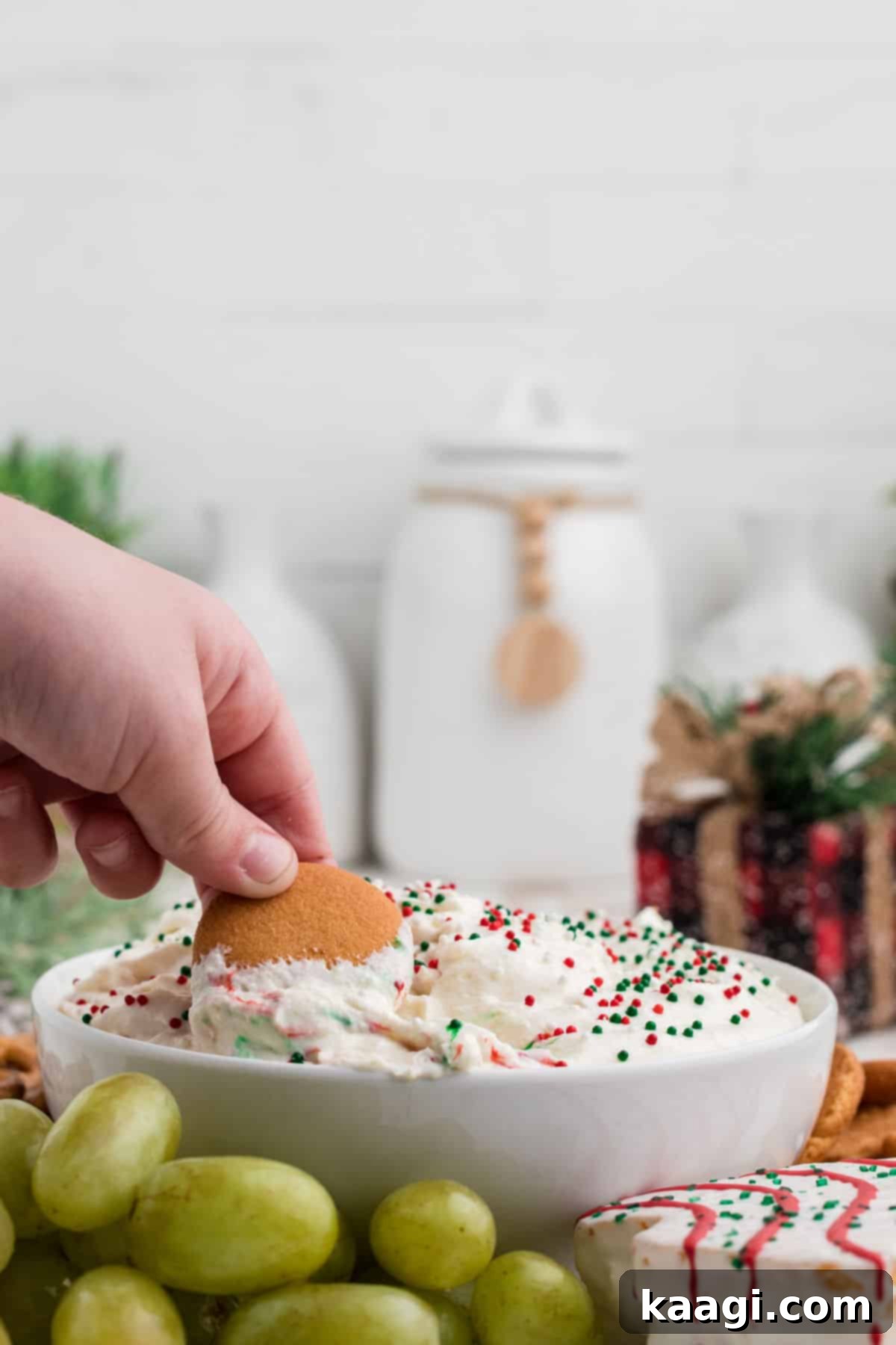 Sparkling Christmas Tree Dip 6 A child's hand dipping a gingerbread cookie into Christmas Tree Dip, surrounded by other dippers like fruit and crackers.