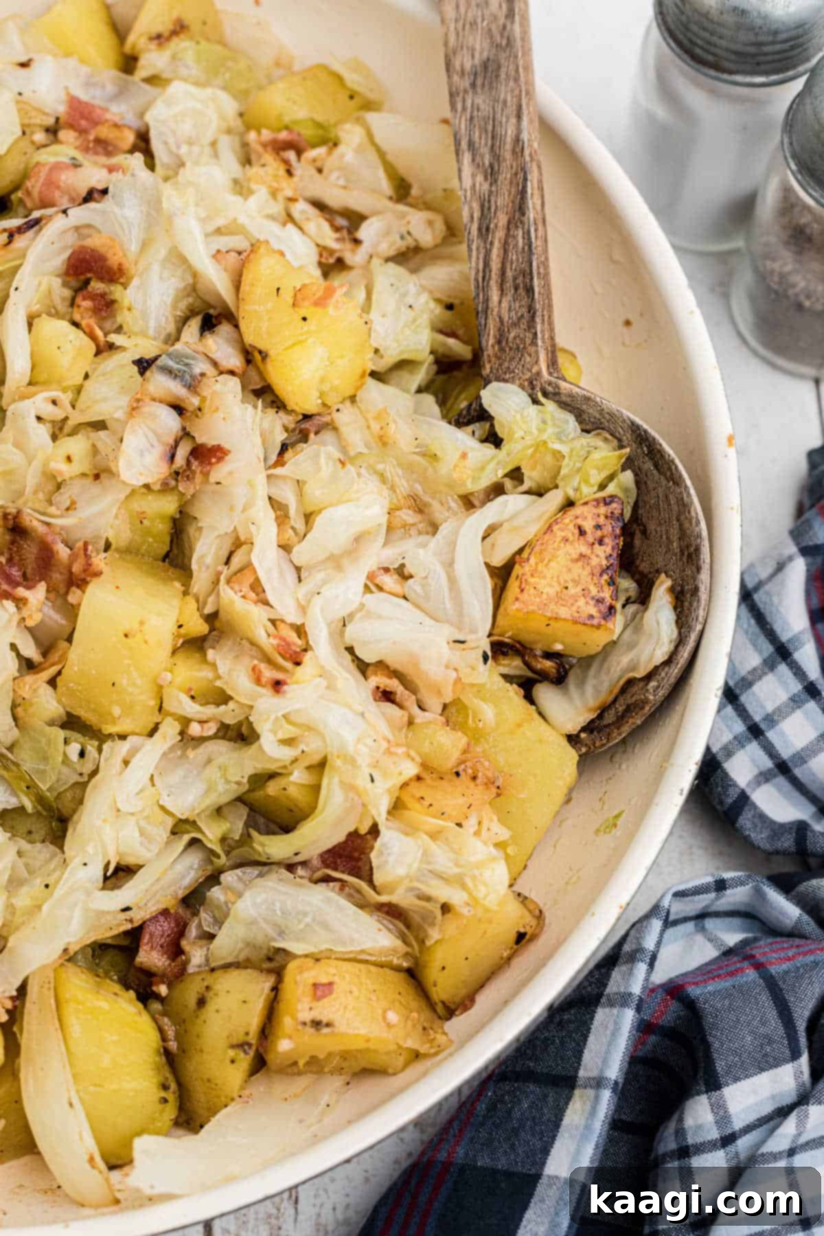 A close-up side view of a skillet filled with golden-brown fried cabbage and potatoes, with a serving spoon scooping a portion.