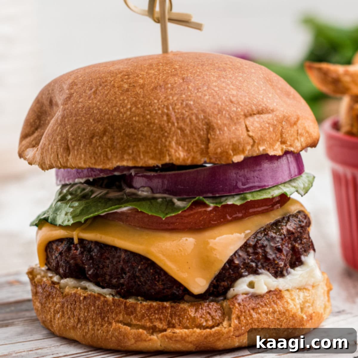 A close-up of a dressed venison burger, garnished with fresh toppings, with a bowl of french fries in the blurred background.